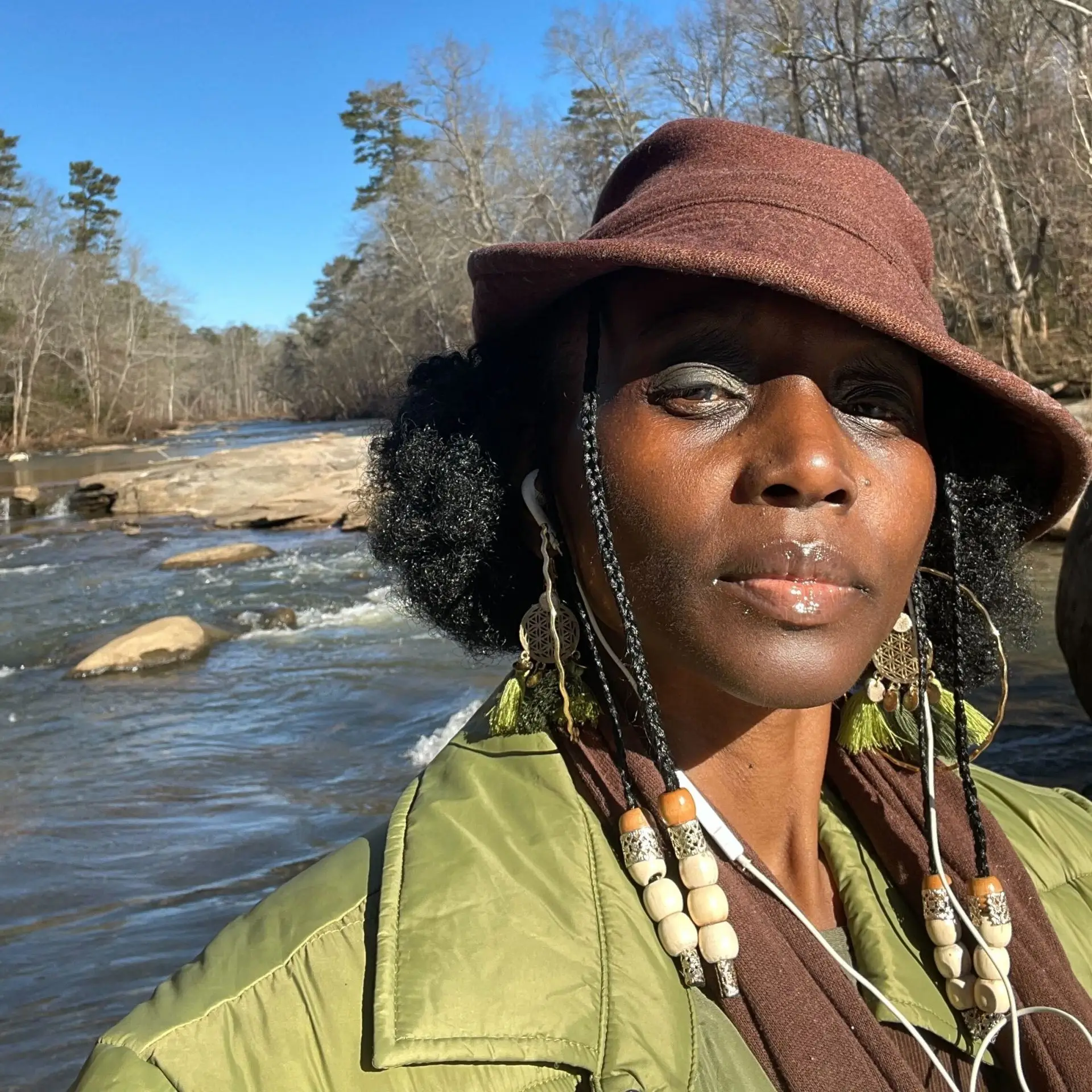 A woman is taking a selfie outdoors near a flowing river. She wears a brown hat, a green jacket, and beaded earrings with braids. Leafless trees line both sides of the river, and the sky is clear and bright blue. Sunlight highlights her face and clothing.